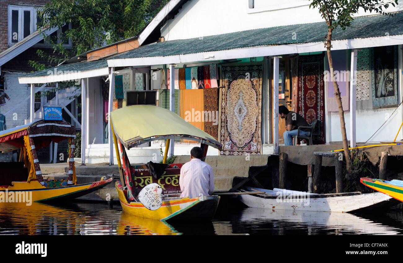 Una shikara, pequeño barco taxi de agua de madera, amarrado por una tienda de alfombras casa en Dal Lago. Dal Lago de Srinagar, Cachemira. Foto de stock