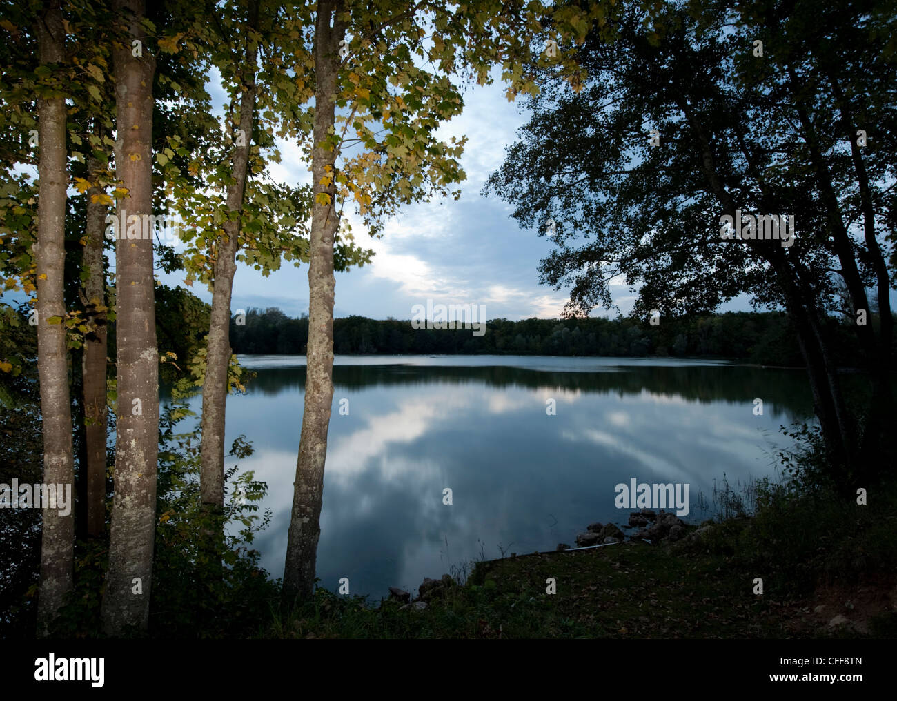 El cielo y las nubes reflejados en el lago en Grez sur Loing al atardecer con árboles en primer plano iluminado con flash Foto de stock