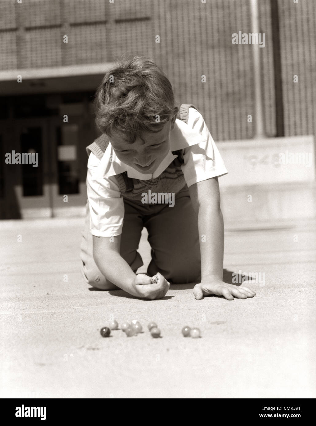 1950 sonriente chico listo para disparar arrodillado en el patio de la escuela TIERRA JUGANDO CANICAS Foto de stock