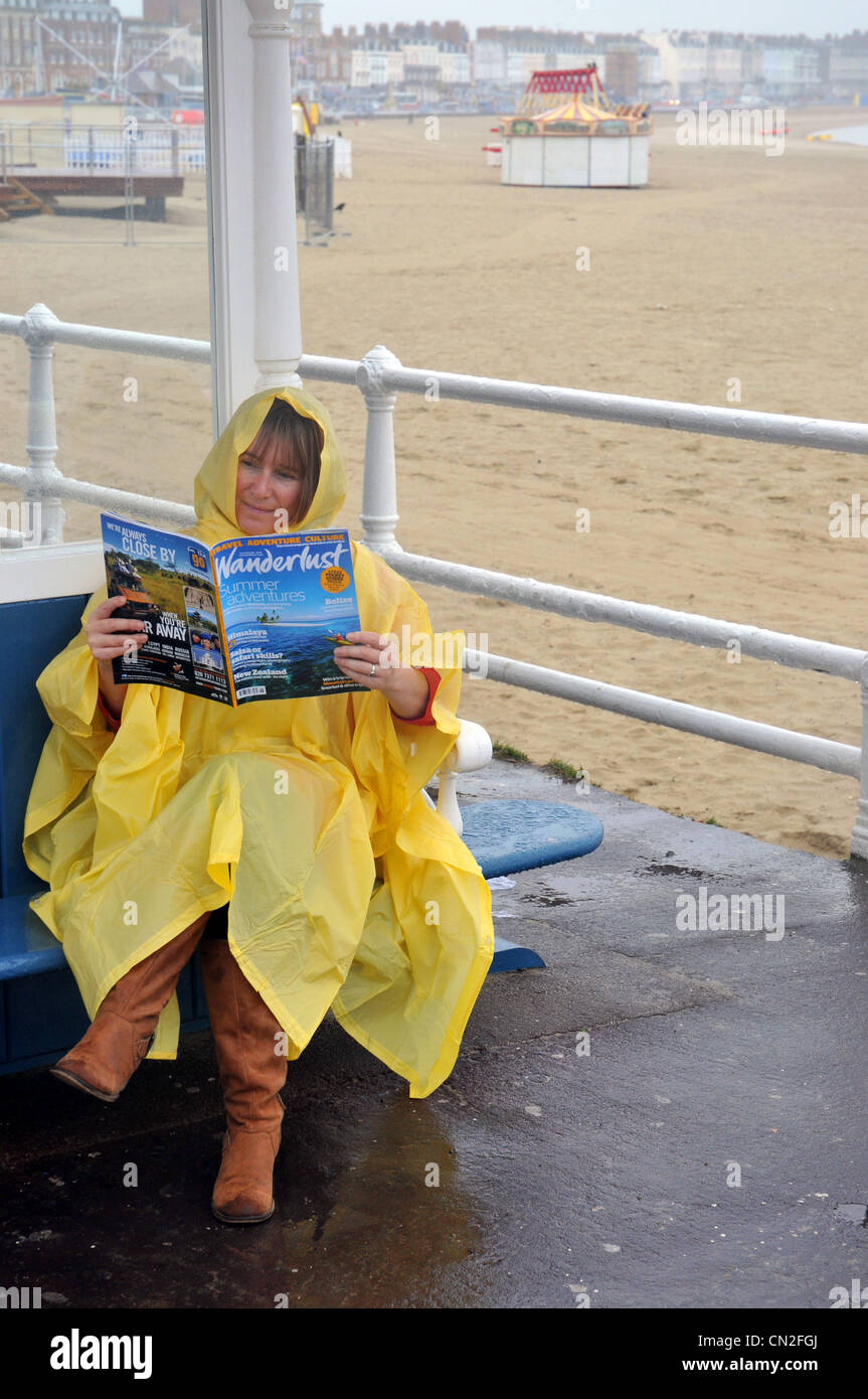 El clima húmedo, mujer lee una revista de vacaciones en un húmedo y frío en la playa de Weymouth Dorset, Inglaterra, Reino Unido Foto de stock