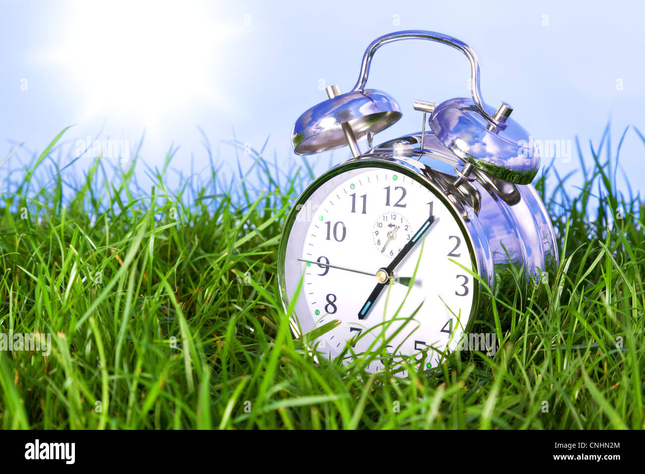 Foto de un reloj despertador de cromo afuera, sentado en la hierba en la mañana de un día soleado. Foto de stock