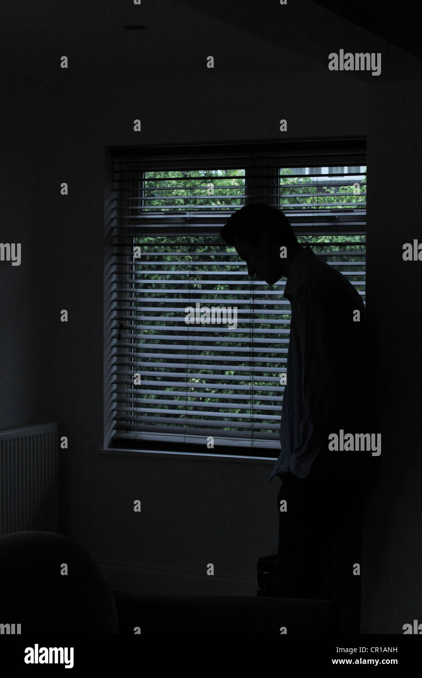 Silueta de un hombre de pie en una habitación oscura con luz de una ventana con persianas mostrando su forma. Foto de stock