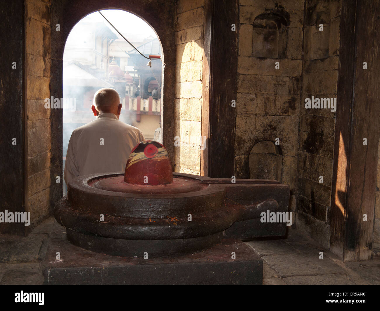 Hombre sentado detrás de un lingam de Shiva, viendo una ceremonia de cremación en el templo Pashupatinath, Katmandú, Nepal, en el sur de Asia Foto de stock
