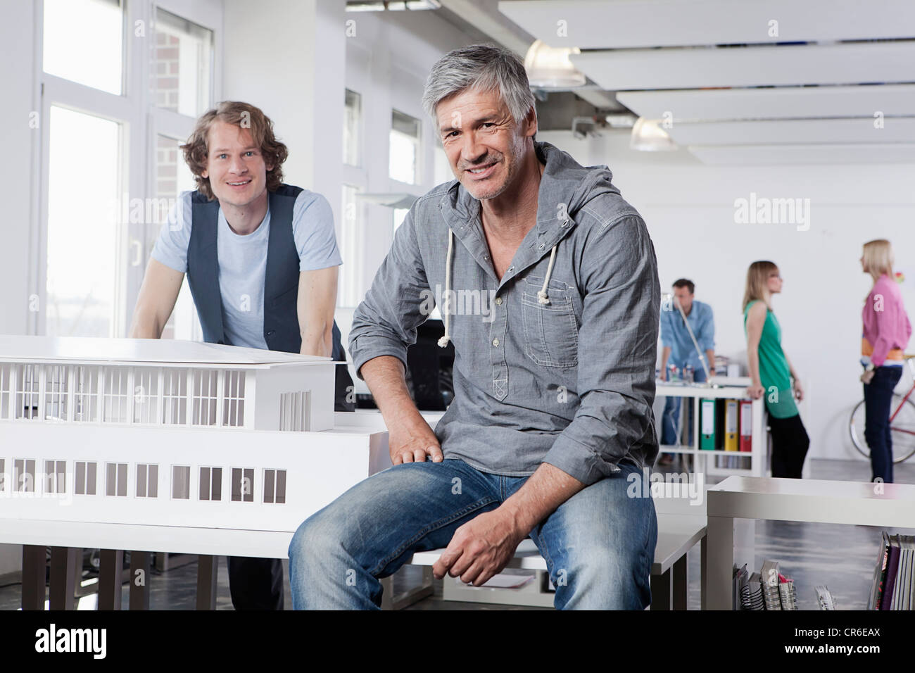 Alemania, Baviera, Munich, hombres con modelo arquitectónico en oficina, compañeros hablando en segundo plano. Foto de stock