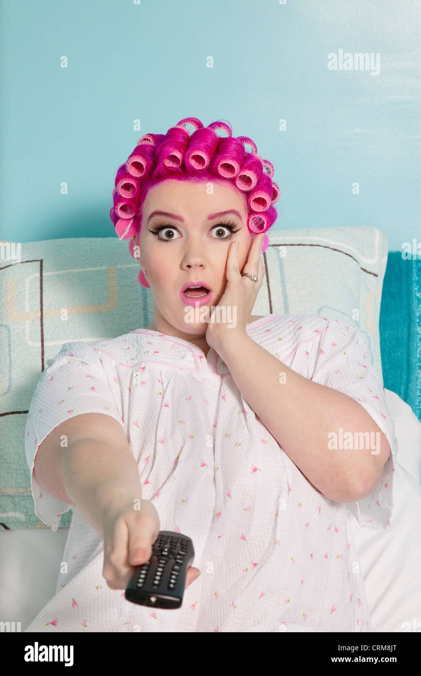 Retrato de mujer sosteniendo conmocionado remoto con rizadores de pelo sentada en la cama Foto de stock