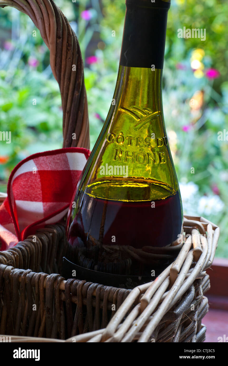 Cotes Du Rhone vino tinto francés botella en cesta de mimbre en tarde de sol con la típica servilleta de picnic y jardín de verano detrás Foto de stock