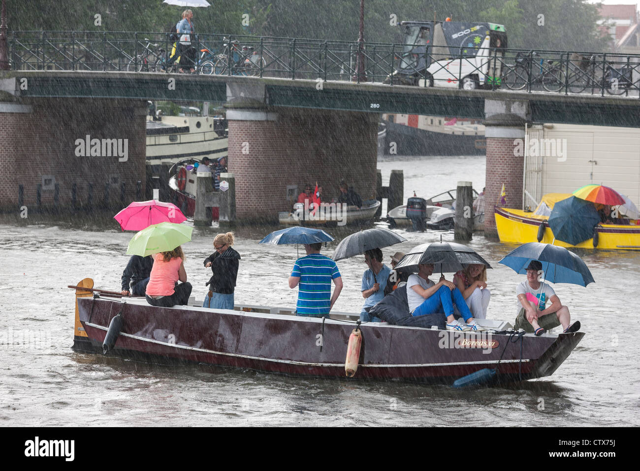 Torrenciales repentinas lluvias de verano en Amsterdam. 9 personas en un pequeño bote, sloop, con sombrillas. Foto de stock