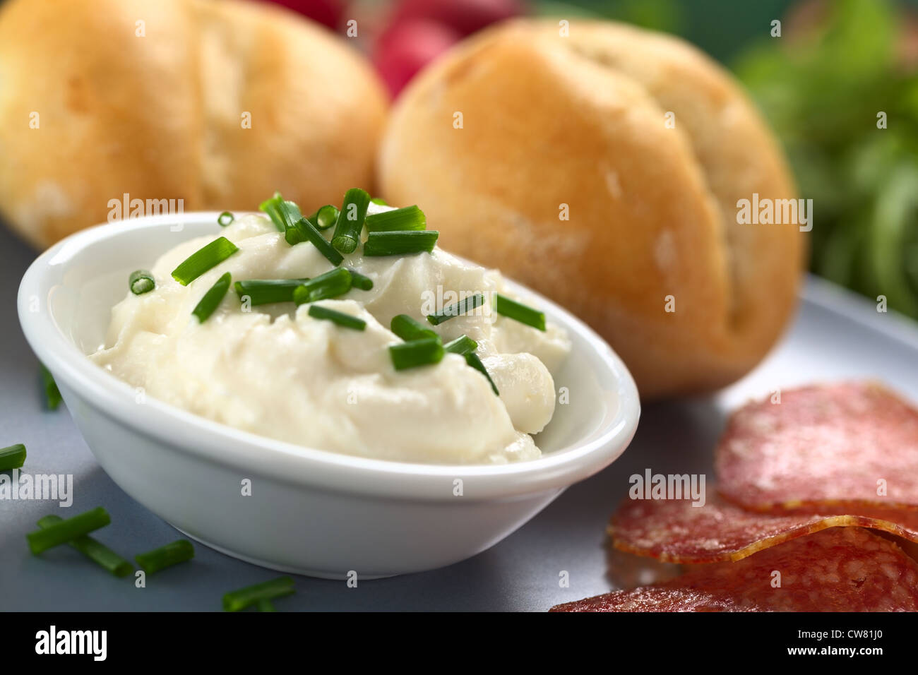 Propagación de crema de queso fresco en un tazón pequeño con cebollinos por encima con rodajas de salami y bollos en la placa Foto de stock