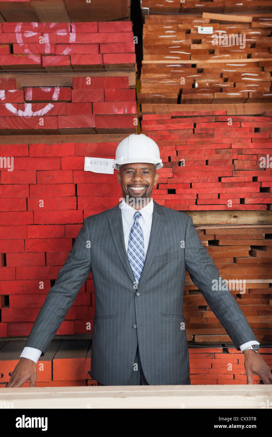 Retrato de feliz contratista macho americano africano con tablones de madera apilados en segundo plano. Foto de stock