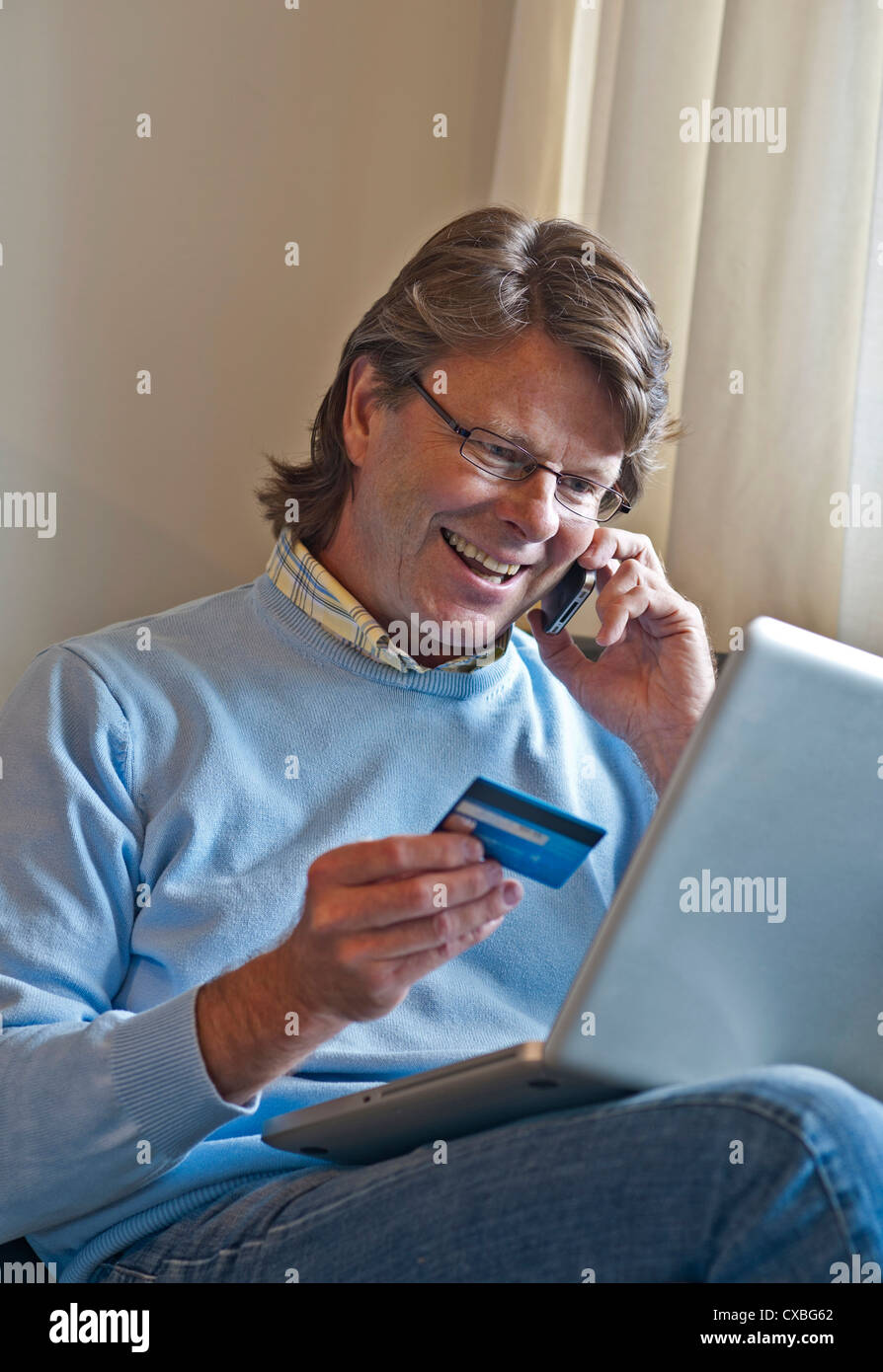 Hombre maduro en casa felizmente haciendo una transacción financiera en línea utilizando su ordenador portátil y un smartphone iPhone 4s Foto de stock