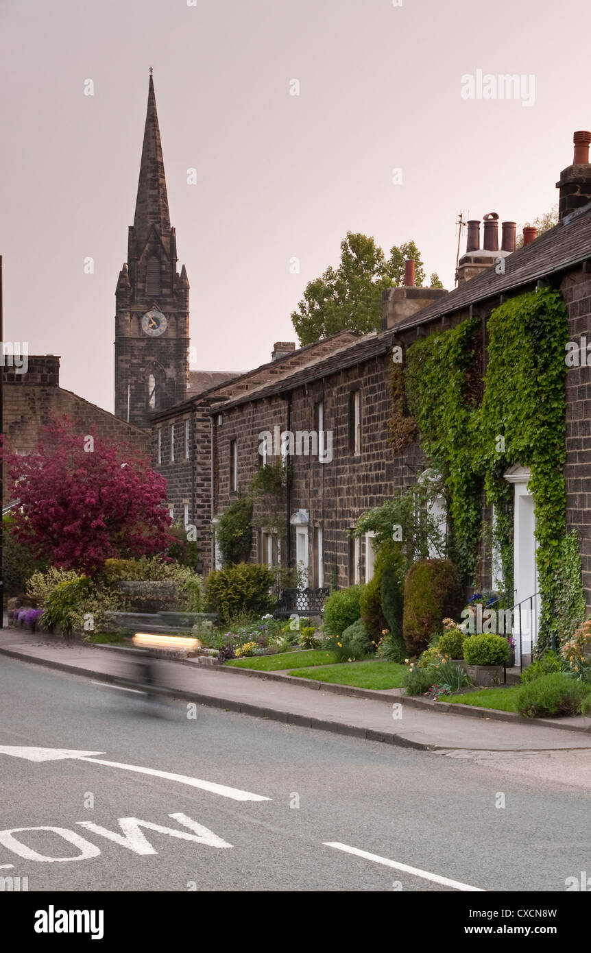 Terraza hilera de casas de carretera construida en piedra con una torre de chapitel y reloj de la Iglesia de Santa María más allá - Burley de Wharfedale, en Yorkshire, Inglaterra, Reino Unido. Foto de stock