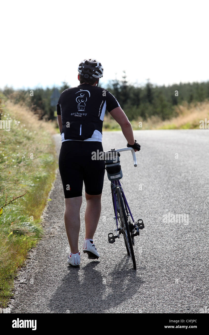 Ciclista camino empujando su bicicleta hasta una colina en Aberdeenshire, Escocia, Reino Unido, en un día soleado, disparos por la espalda con vista posterior mostrando Foto de stock