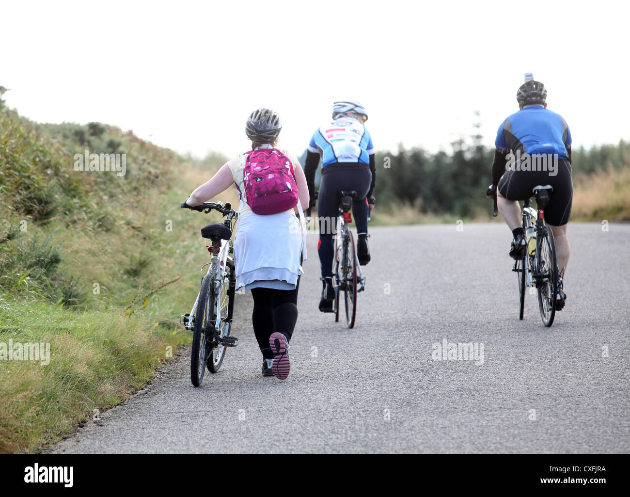 Hembra ciclista de carretera empujando su bicicleta hasta una colina en Aberdeenshire, Escocia, Reino Unido, como dos hombres pasado ciclo Foto de stock