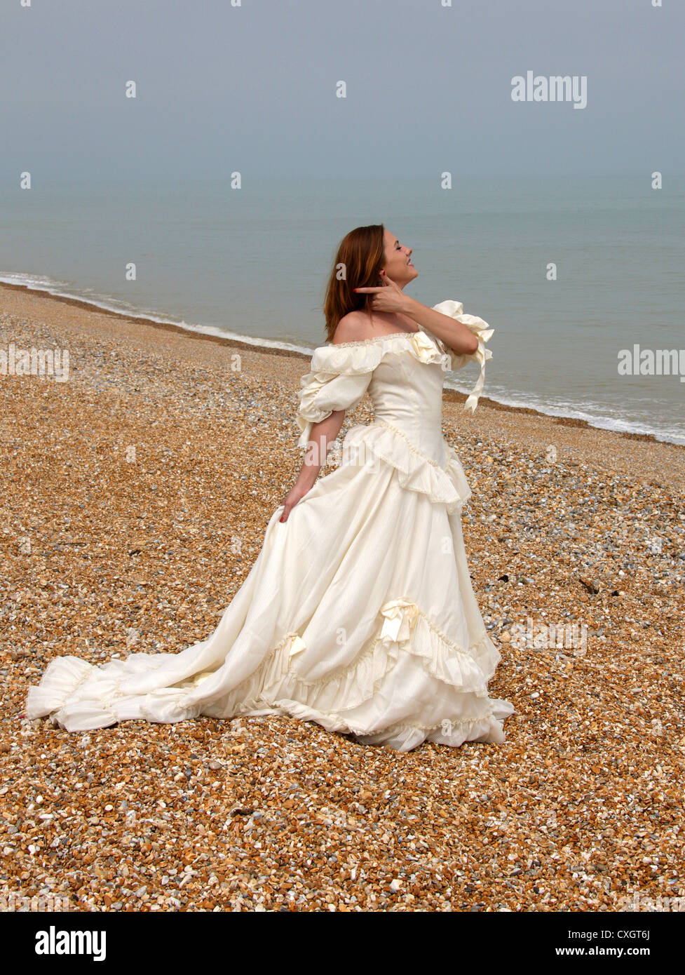 Chica que llevaba un vestido blanco crema en una playa de guijarros por el mar. Foto de stock
