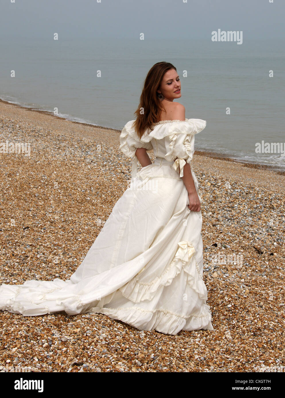 Chica que llevaba un vestido blanco crema en una playa de guijarros por el mar. Foto de stock