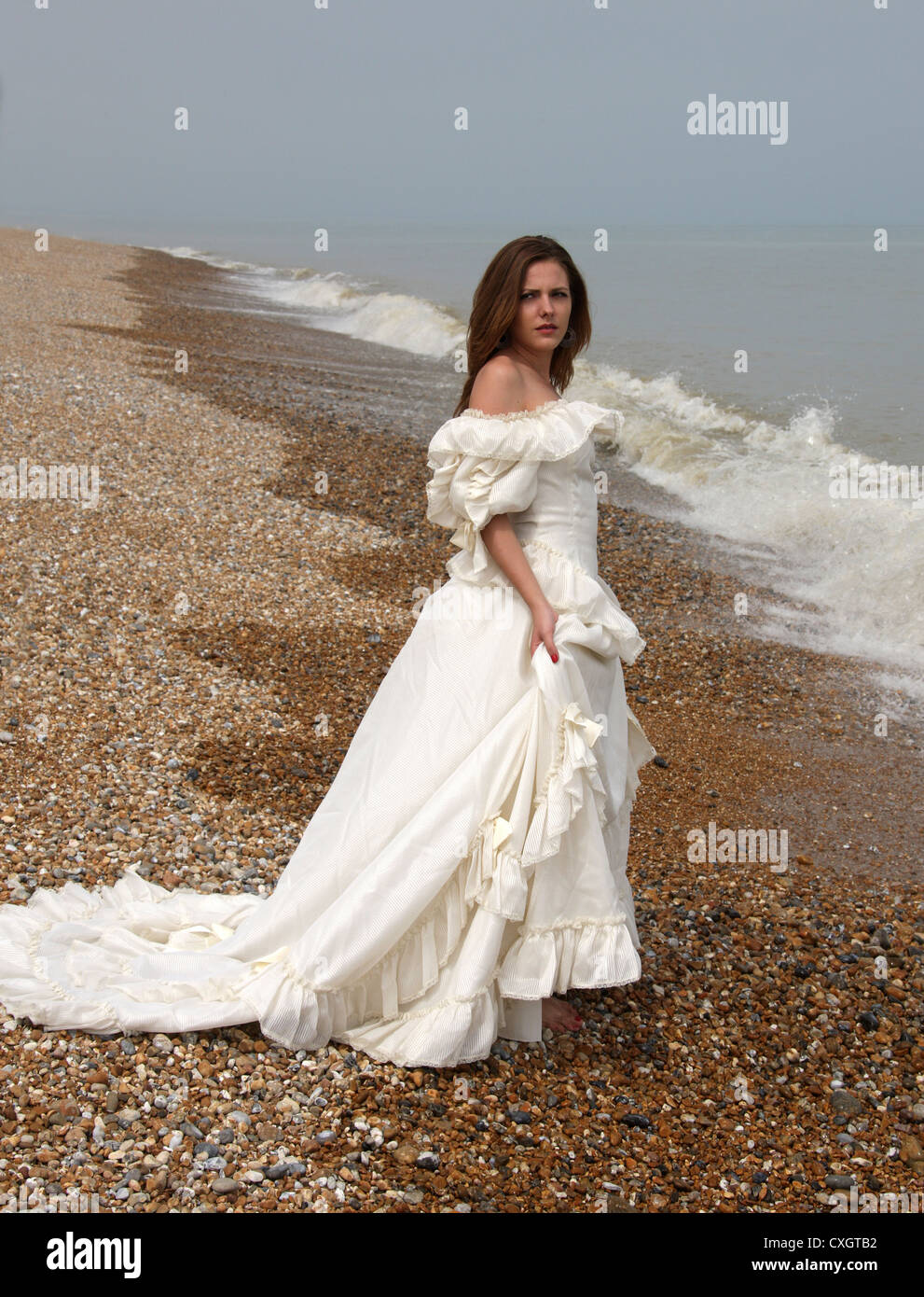 Chica que llevaba un vestido blanco crema en una playa de guijarros por el mar. Foto de stock