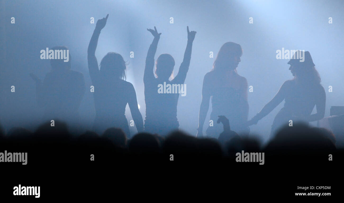 Ambiente durante un concierto de rock en directo con siluetas de mujeres bailando en el escenario con rockeros brazos arriba en el aire Foto de stock