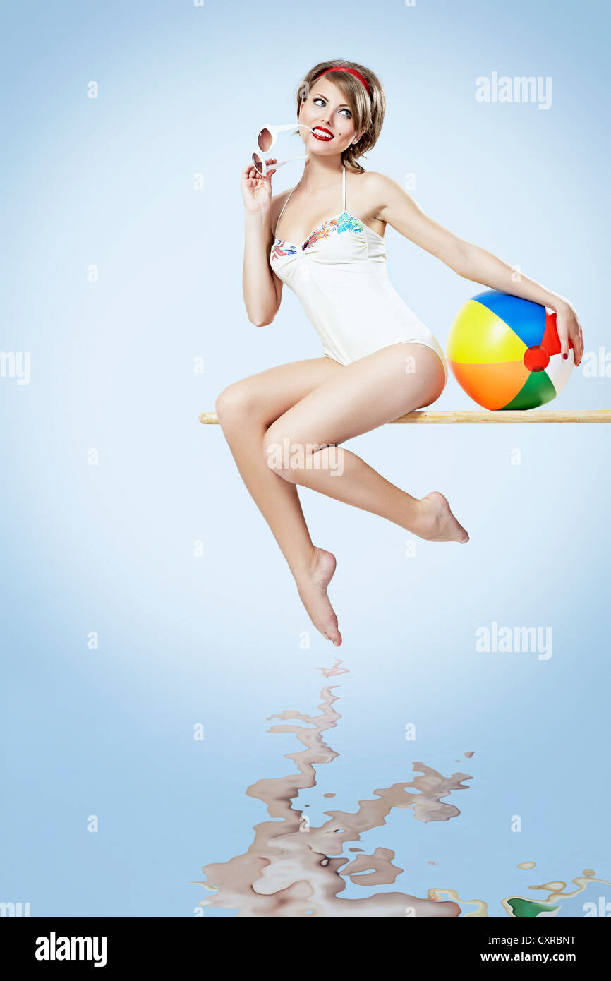 Mujer joven vistiendo un traje de baño blanco sentado en un trampolín y sosteniendo un balón de playa, pin-up Foto de stock
