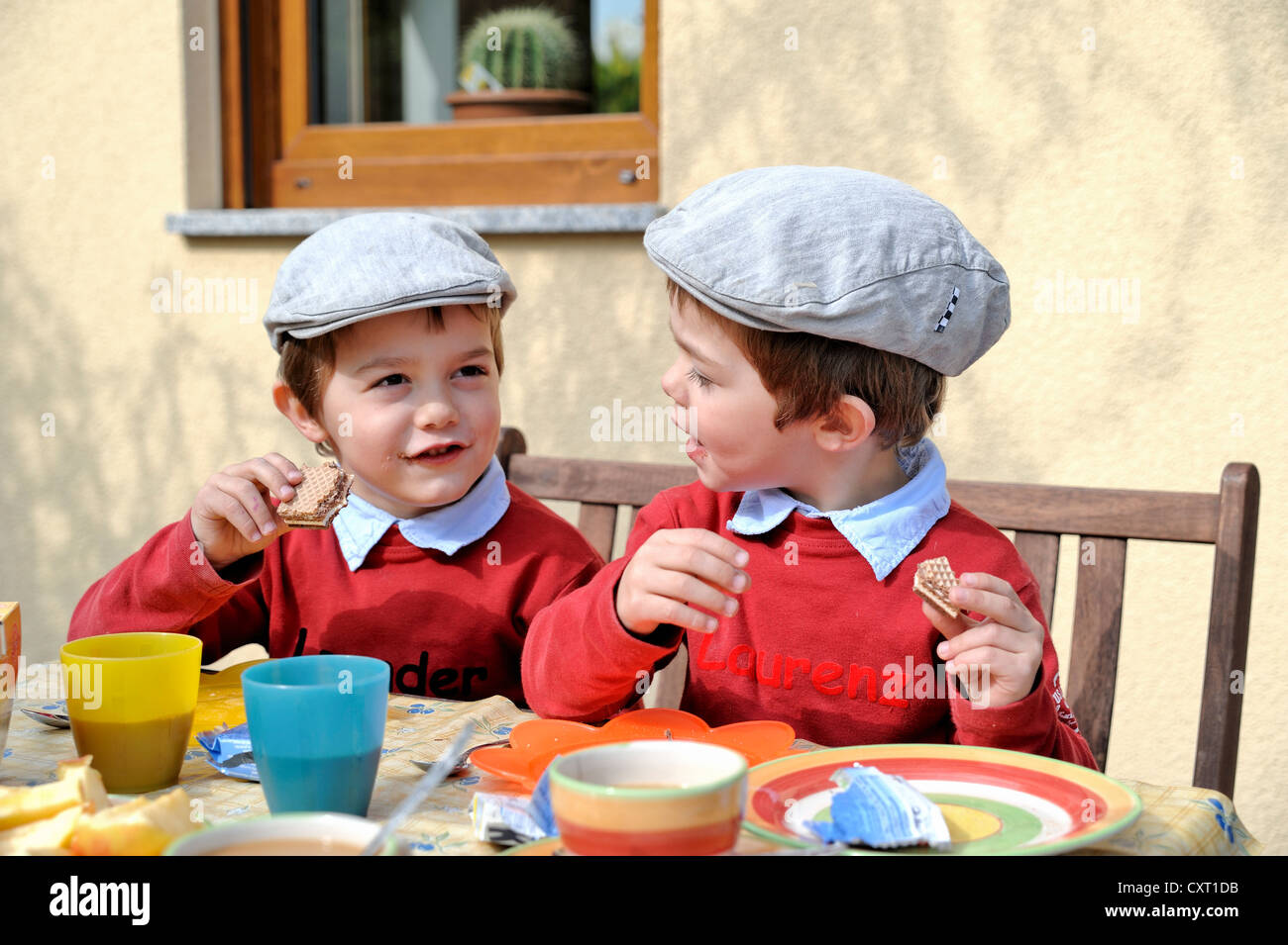Gemelos, 4, vistiendo gorras planas, sentarse y comer en una mesa Foto de stock