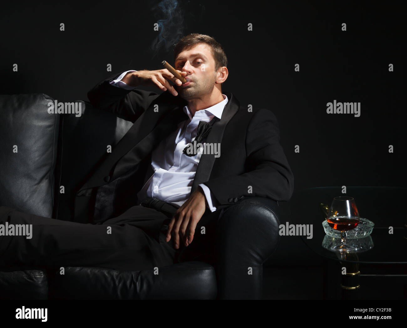 Hombre macho en un elegante esmoquin sentado en la oscuridad de una discoteca puffing sobre un cigarro y beber brandy o coñac Foto de stock