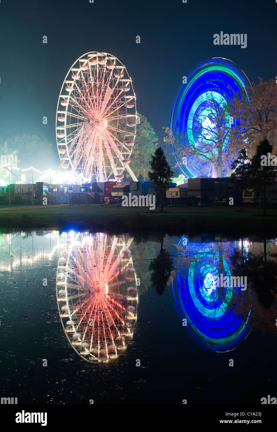 Las luces de dos ruedas de ferris en un parque de diversiones se refleja en el agua de un estanque Foto de stock