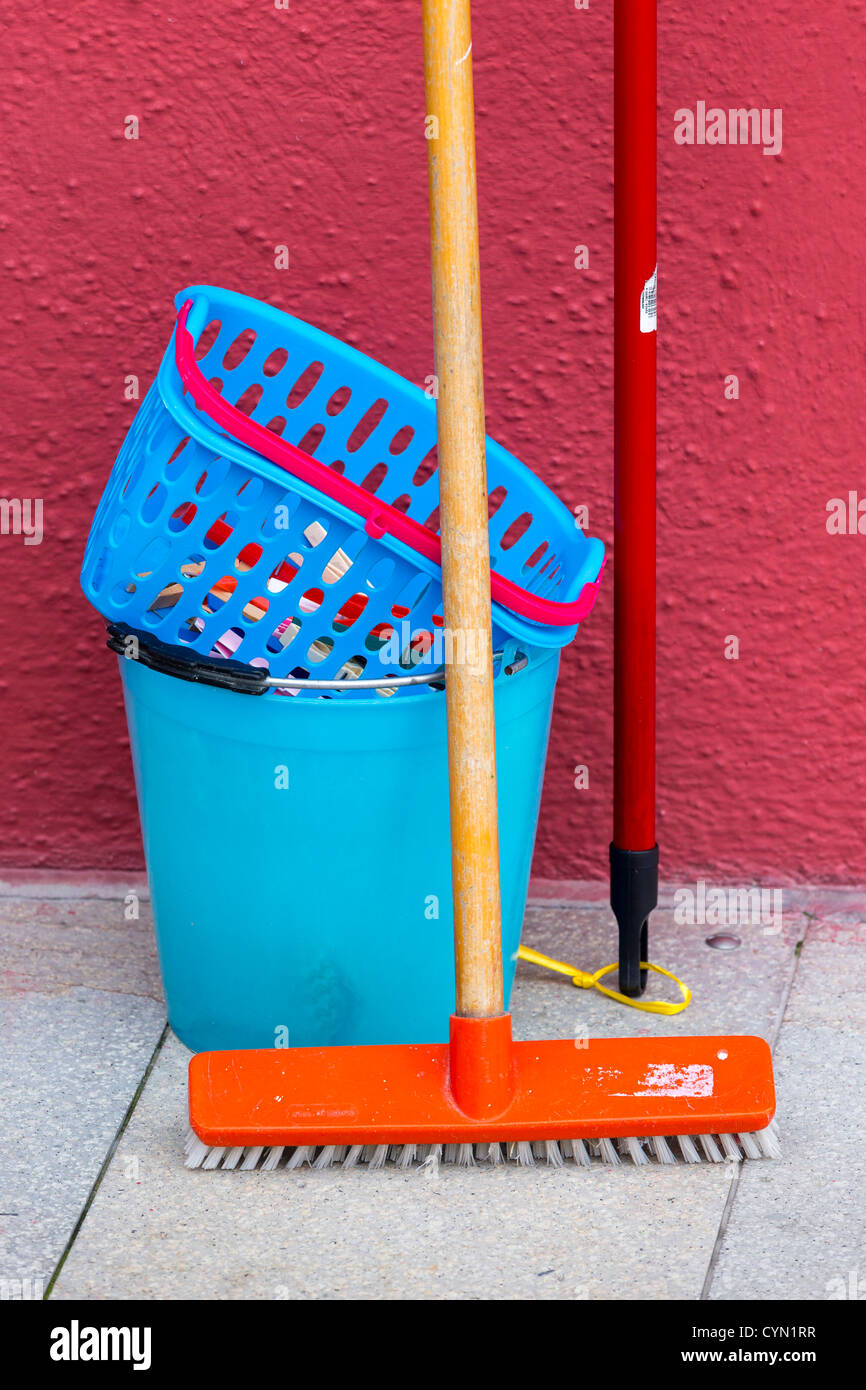 Cuchara de plástico azul y cesto de ropa de color naranja con un cepillo para el suelo contra una pared roja Foto de stock