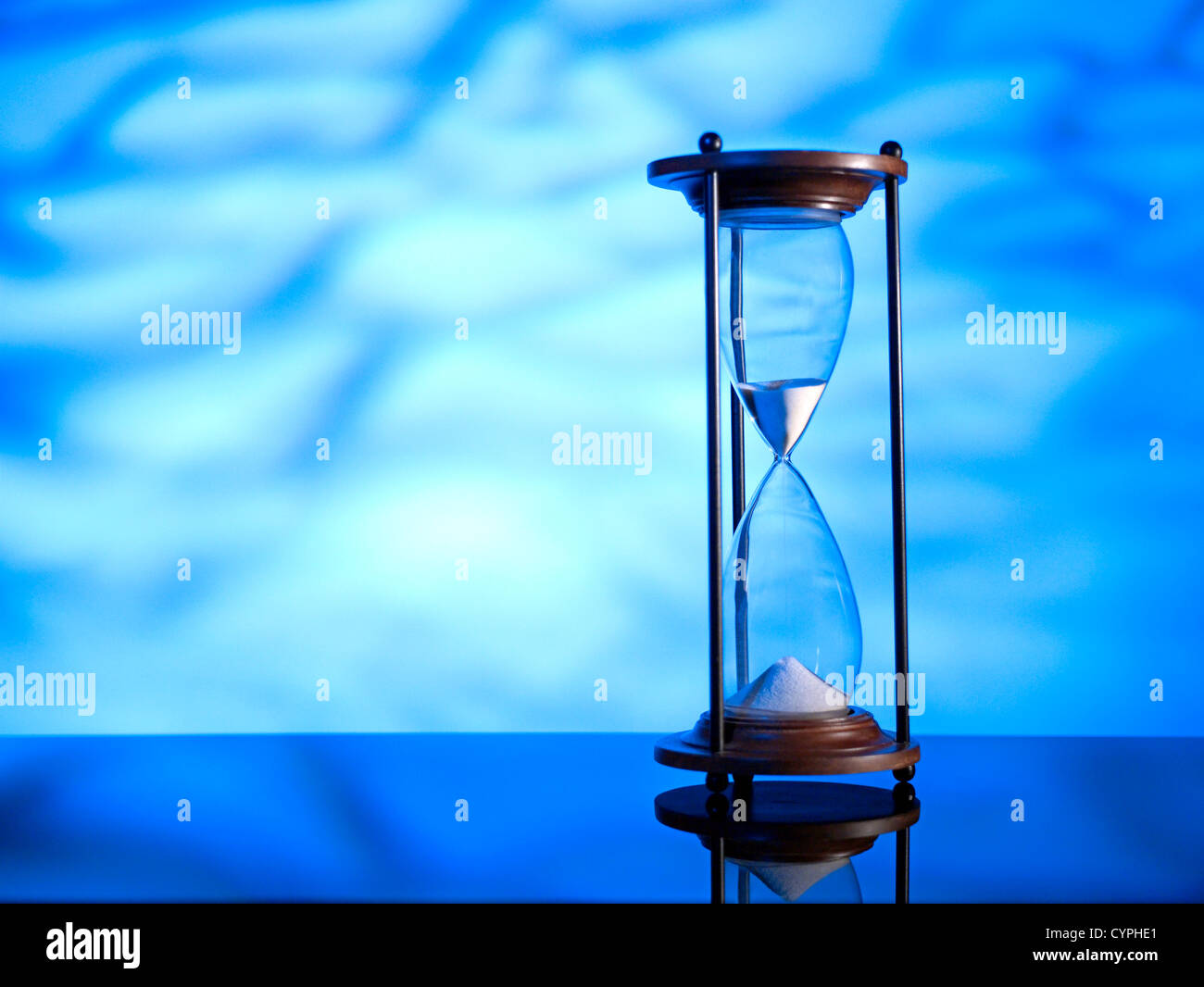 Un reloj de arena de cristal clásico con arena que fluye hacia abajo se encuentra prominentemente en el foco contra un telón de fondo azul vivo, reflejando el concepto de pasar el tiempo. Foto de stock