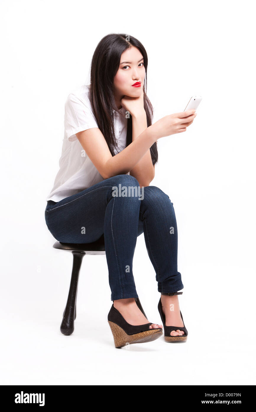 Retrato de hermosa mujer joven en casuals sentado en taburete con teléfono móvil contra el fondo blanco. Foto de stock