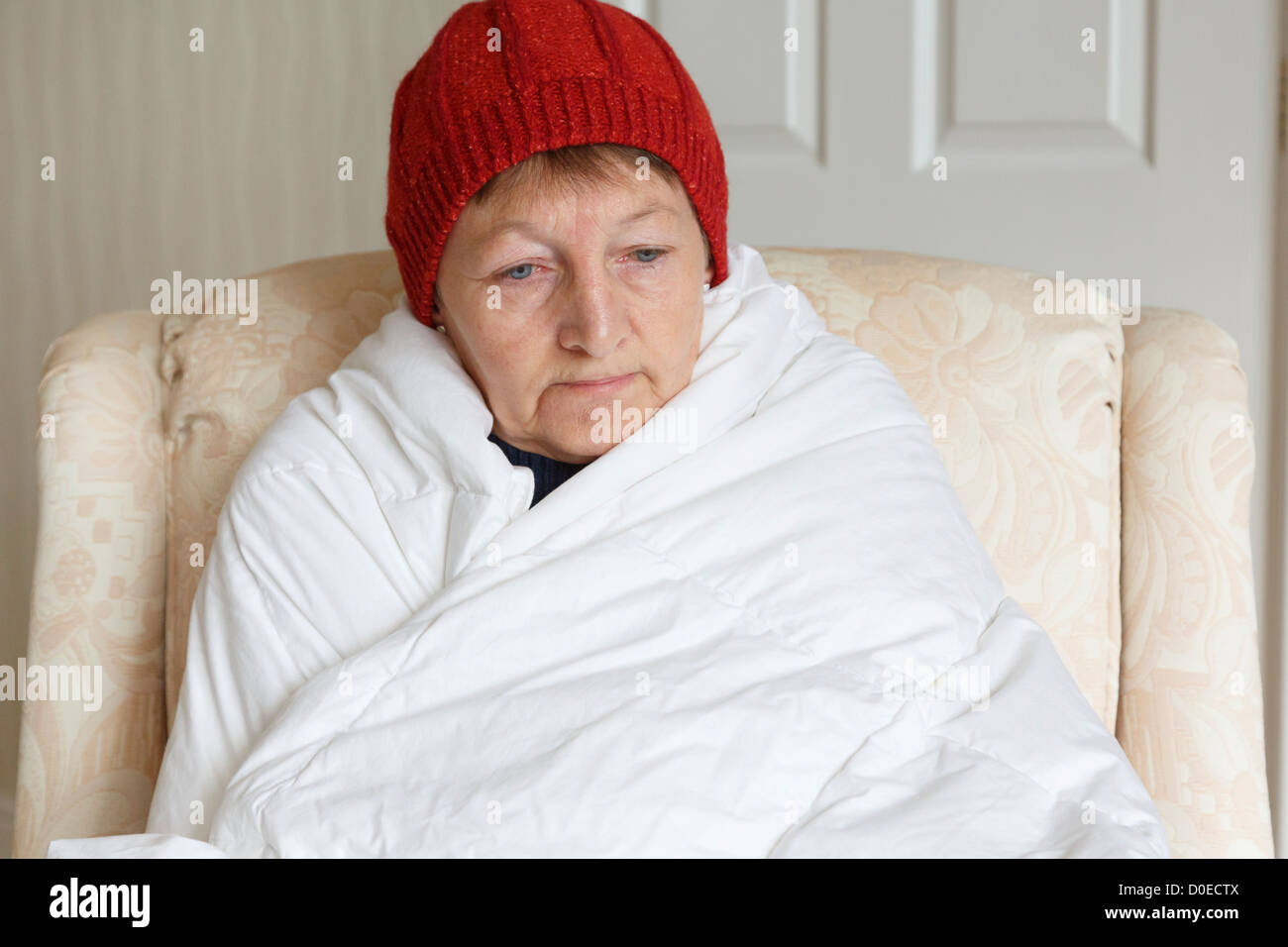 Mujer mayor sensación de frío deprimida y sola, con sombrero y envuelta en un edredón intentando mantener en casa cálida y acogedora en invierno. Inglaterra Gran Bretaña Foto de stock