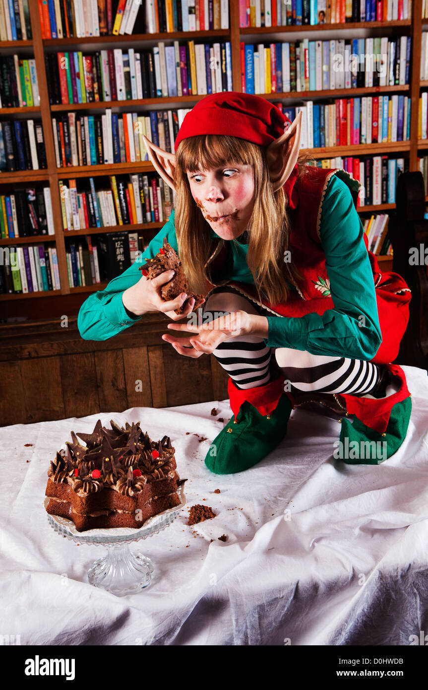 Elf personaje vestido con traje rojo y verde Navidad comiendo una tarta de chocolate en una librería o biblioteca. Foto de stock