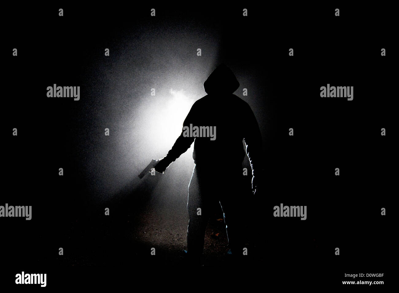 Noche neblinosa Tirador: Una silueta oscura de un hombre encapuchado sosteniendo una pistola semiautomática a su lado de una manera amenazadora. Foto de stock