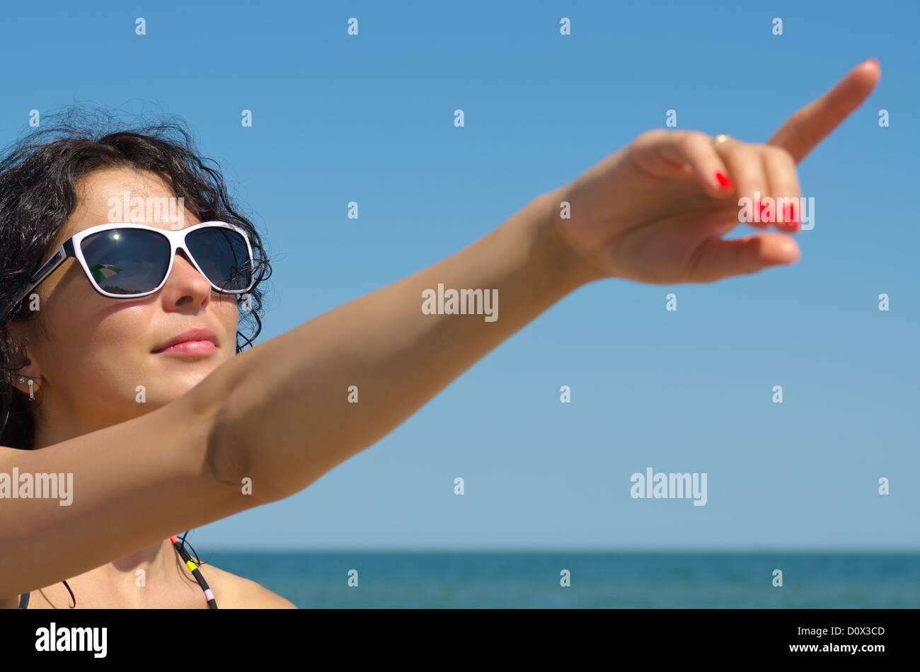 Bella mujer en gafas de sol con un brazo extendido apuntando al cielo azul por encima de su cabeza Foto de stock