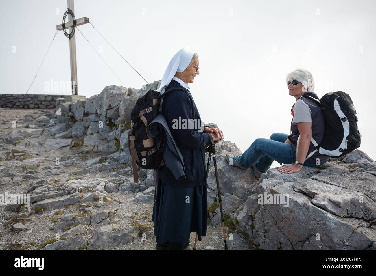 Una monja de senderismo chats con un compañero caminante junto a una cruz en las nubes en el Hafelekar pico en los Alpes por encima de Innsbruck. Foto de stock