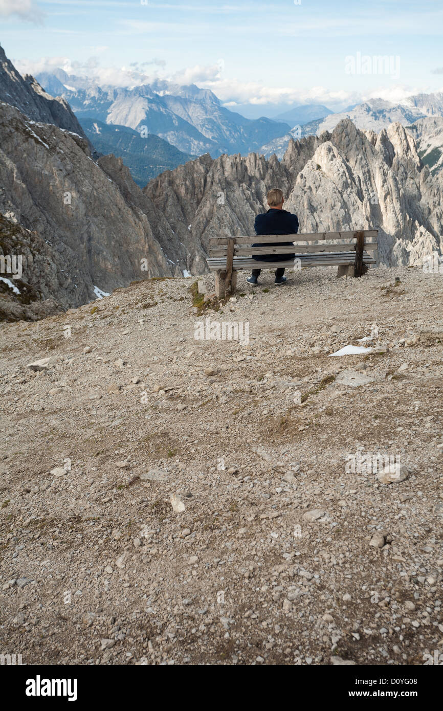 Hombre solitario se sienta en el borde de la banqueta en el pico de la montaña austriaca, mirando a través de los Alpes con capas de escarpadas montañas rocosas. Foto de stock