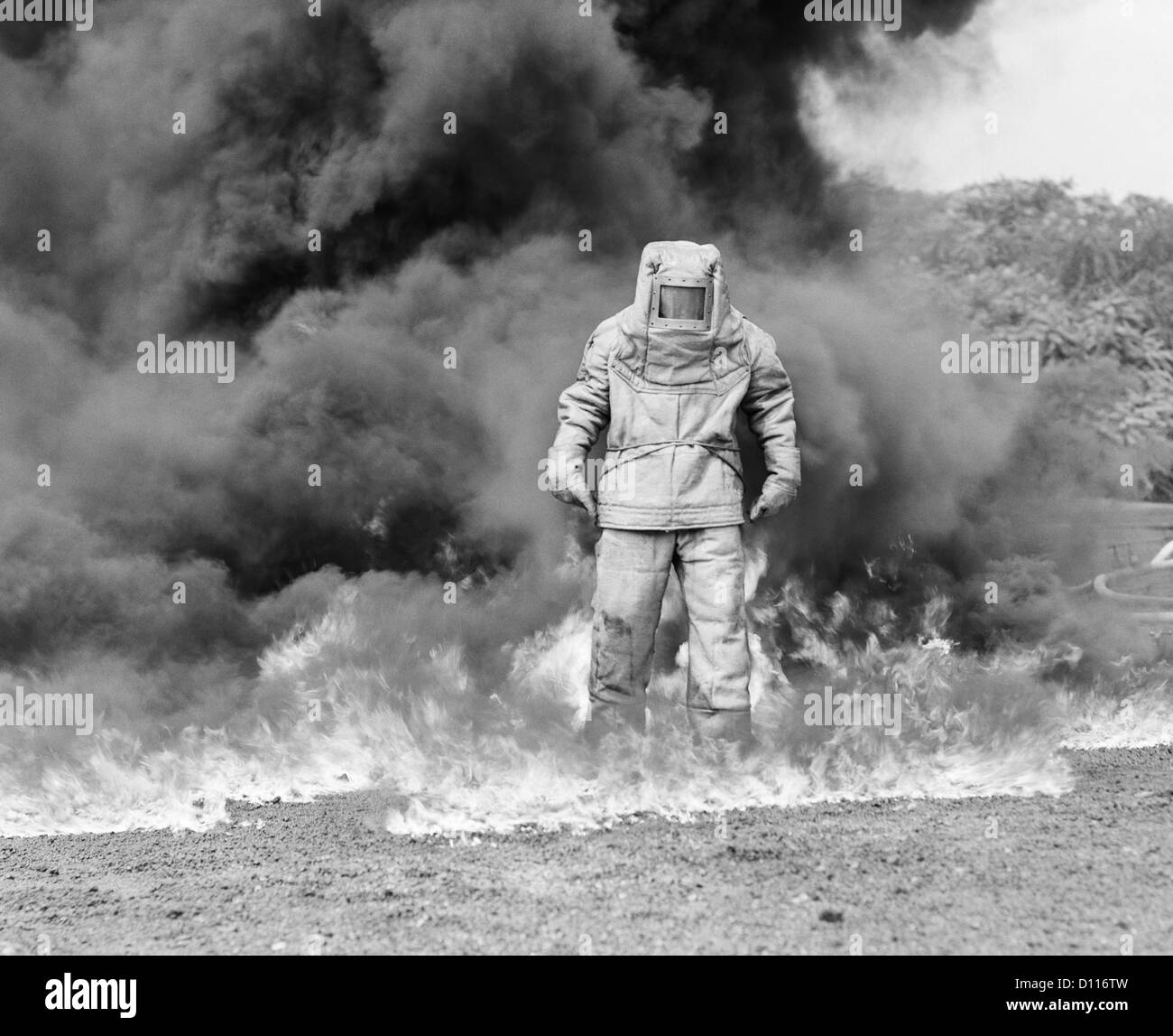 1960 bombero en traje de amianto en pie en humo y llamas Foto de stock