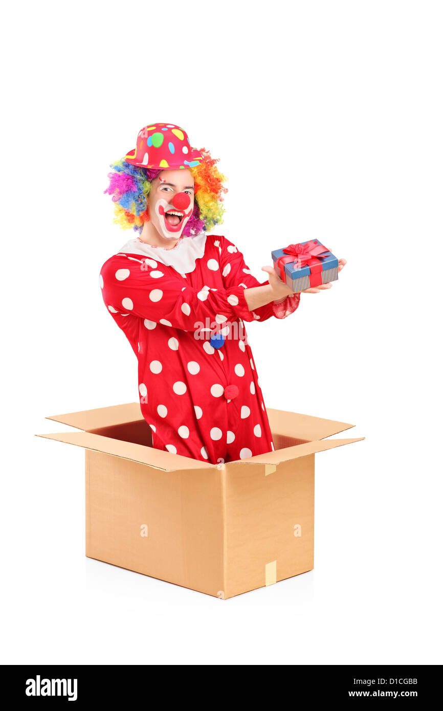 Payaso sonriente en una caja de cartón sosteniendo un regalo aislado sobre fondo blanco. Foto de stock