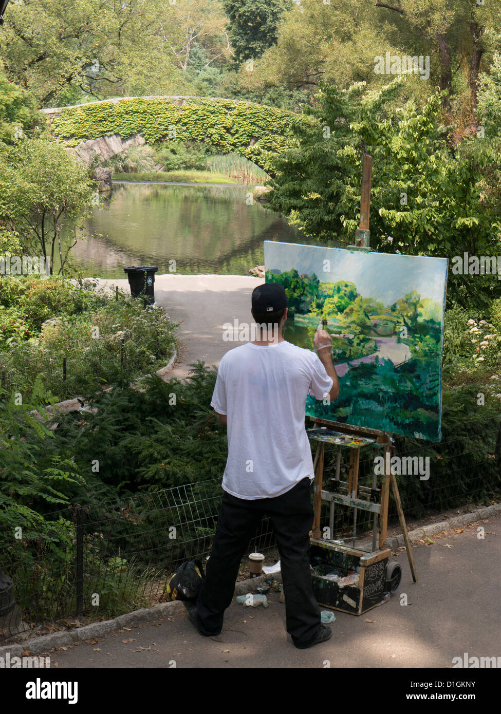 Artista con caballete Pintura y pinta la pacífica escena ante él de los árboles y del agua con puente en Central Park, NYC Foto de stock