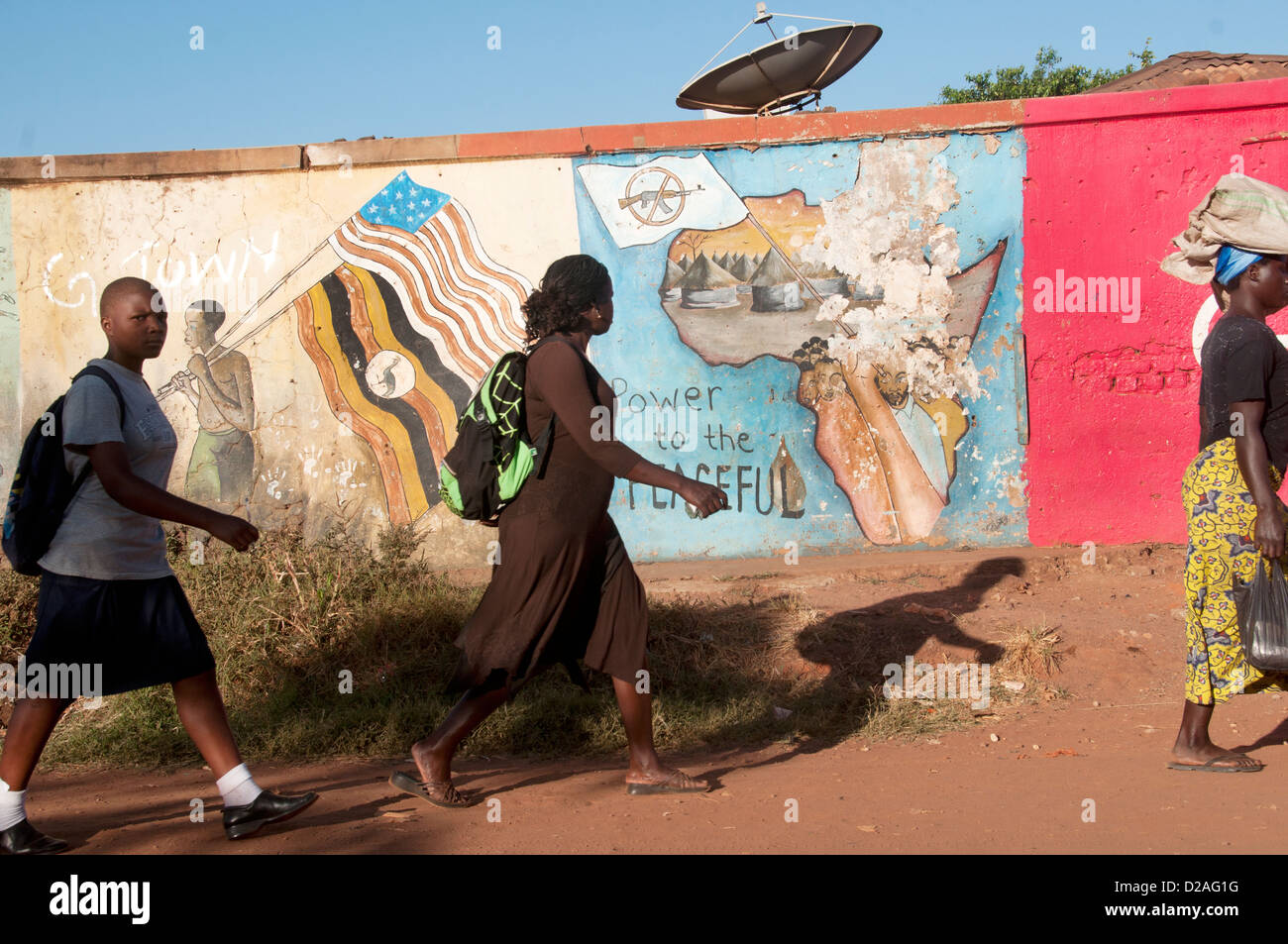 El norte de Uganda, dos mujeres y una colegiala camina más allá de una pared pintada con un mural que muestra un mapa de África en las primeras horas de la mañana el sol Foto de stock
