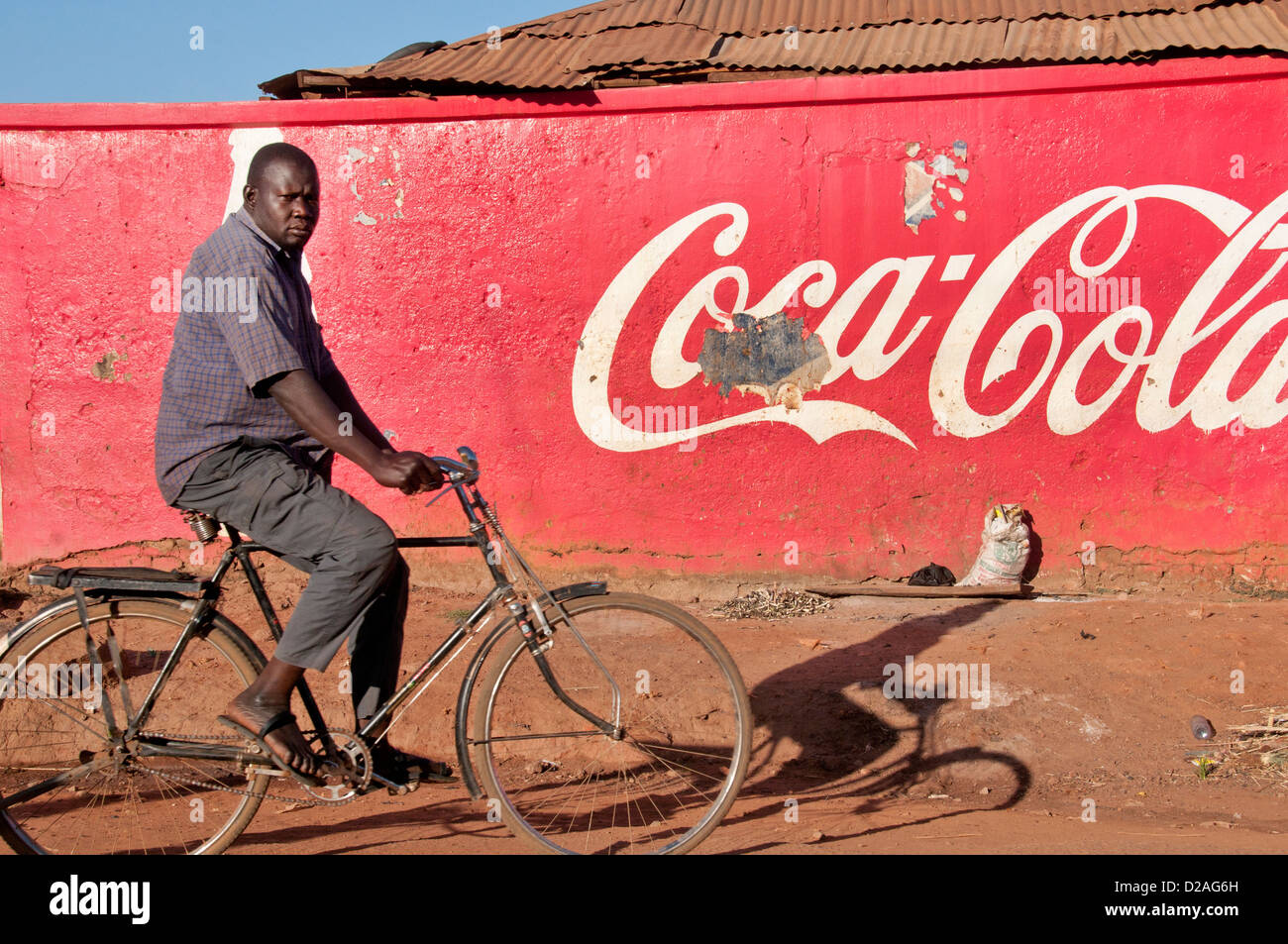El norte de Uganda. Un hombre en una bicicleta ciclos pasados una pared pintada con un anuncio de Coca Cola en las primeras horas de la mañana el sol Foto de stock