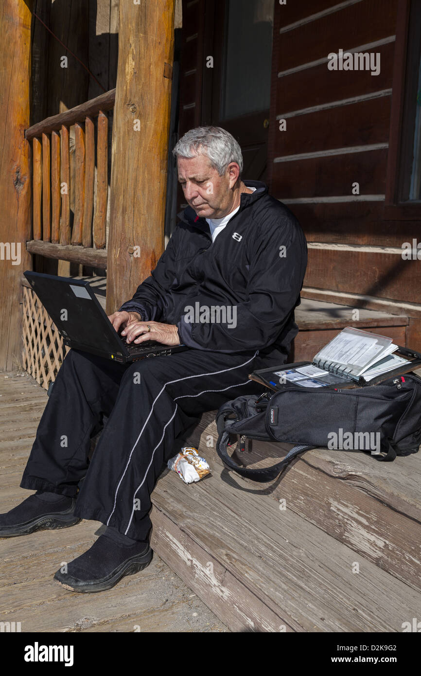 Hombre sentado en un escalón de madera usando el hotspot local creando así su propia oficina al aire libre en un ordenador portátil en el centro de Dubois, Wyoming EE.UU Foto de stock