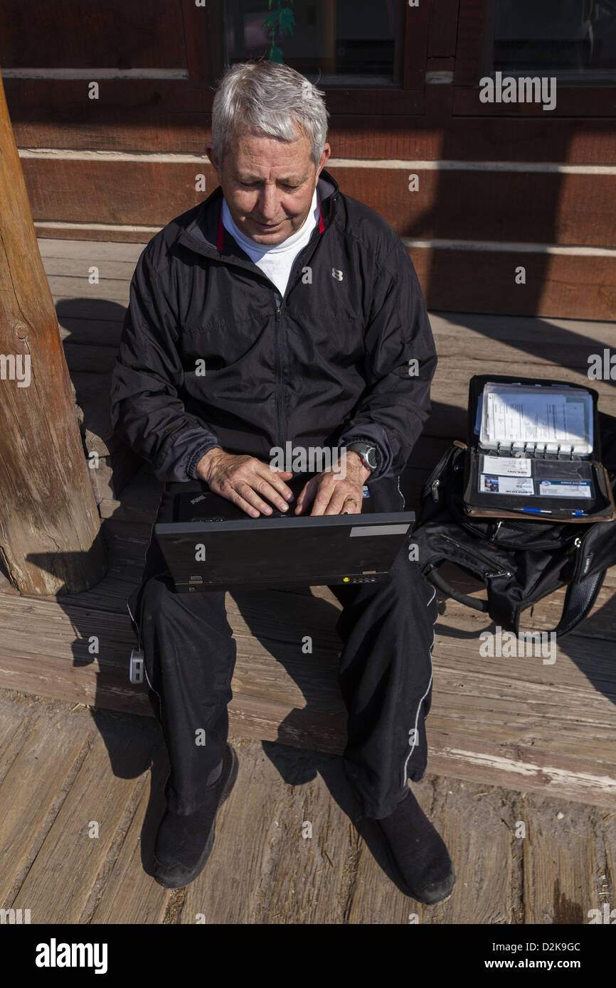 Senior Man sentado en un escalón de madera utilizando el punto de acceso WiFi local, creando así su propia oficina al aire libre en el centro de Dubois, Wyoming EE.UU.. Foto de stock