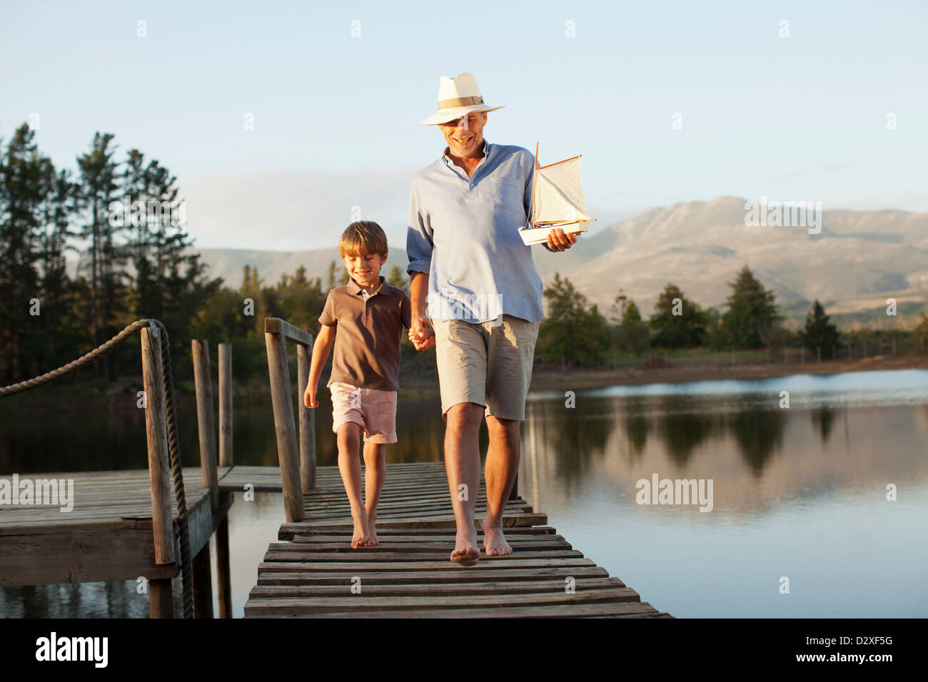 Sonriente, abuelo y nieto de juguete con velero tomados de la mano y caminar a lo largo de muelle sobre el lago Foto de stock