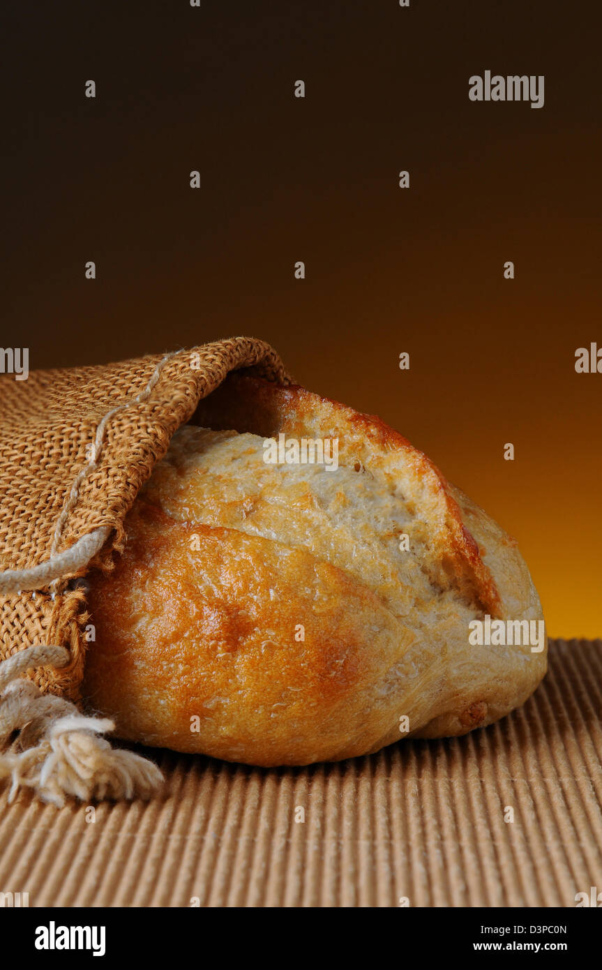 Primer plano de una hogaza de pan en un saco de arpillera en una superficie ondulada y una cálida luz de claro a oscuro. Foto de stock