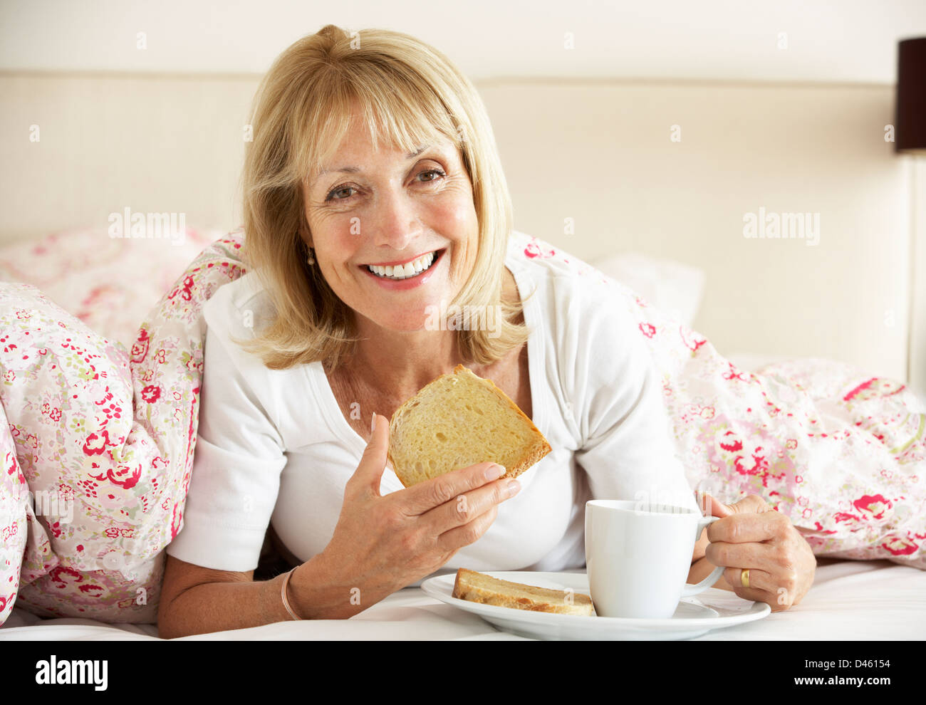 Mujer Senior acurrucada bajo el edredón desayunando Foto de stock