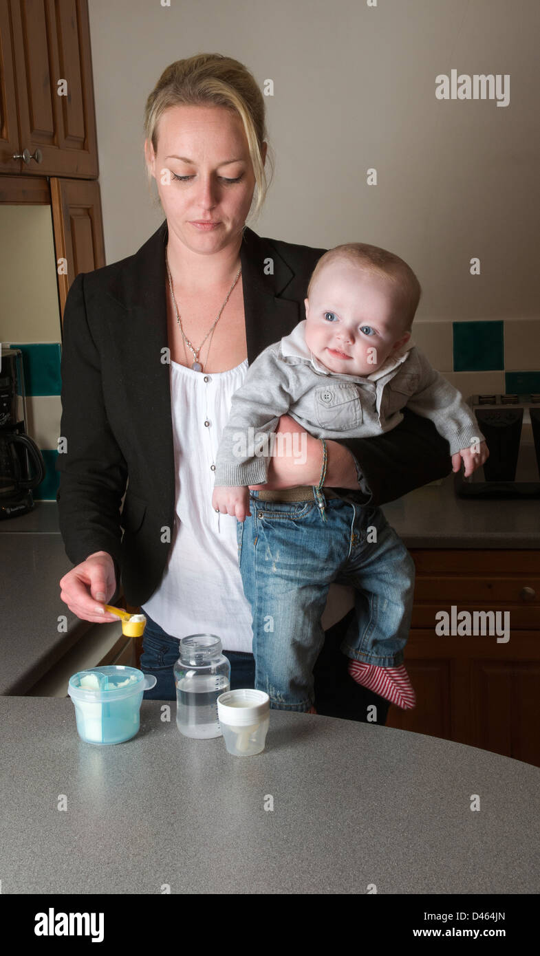 La joven madre la celebración de Baby Boy y preparar su leche beber de pie en la cocina Foto de stock