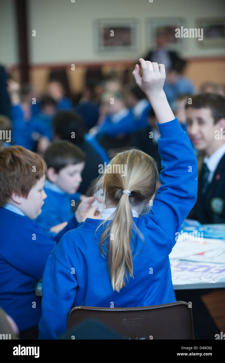 Alumno de la escuela junior en un aula con su mano en el aire contestando a una pregunta del profesor, vestido con un uniforme azul Foto de stock