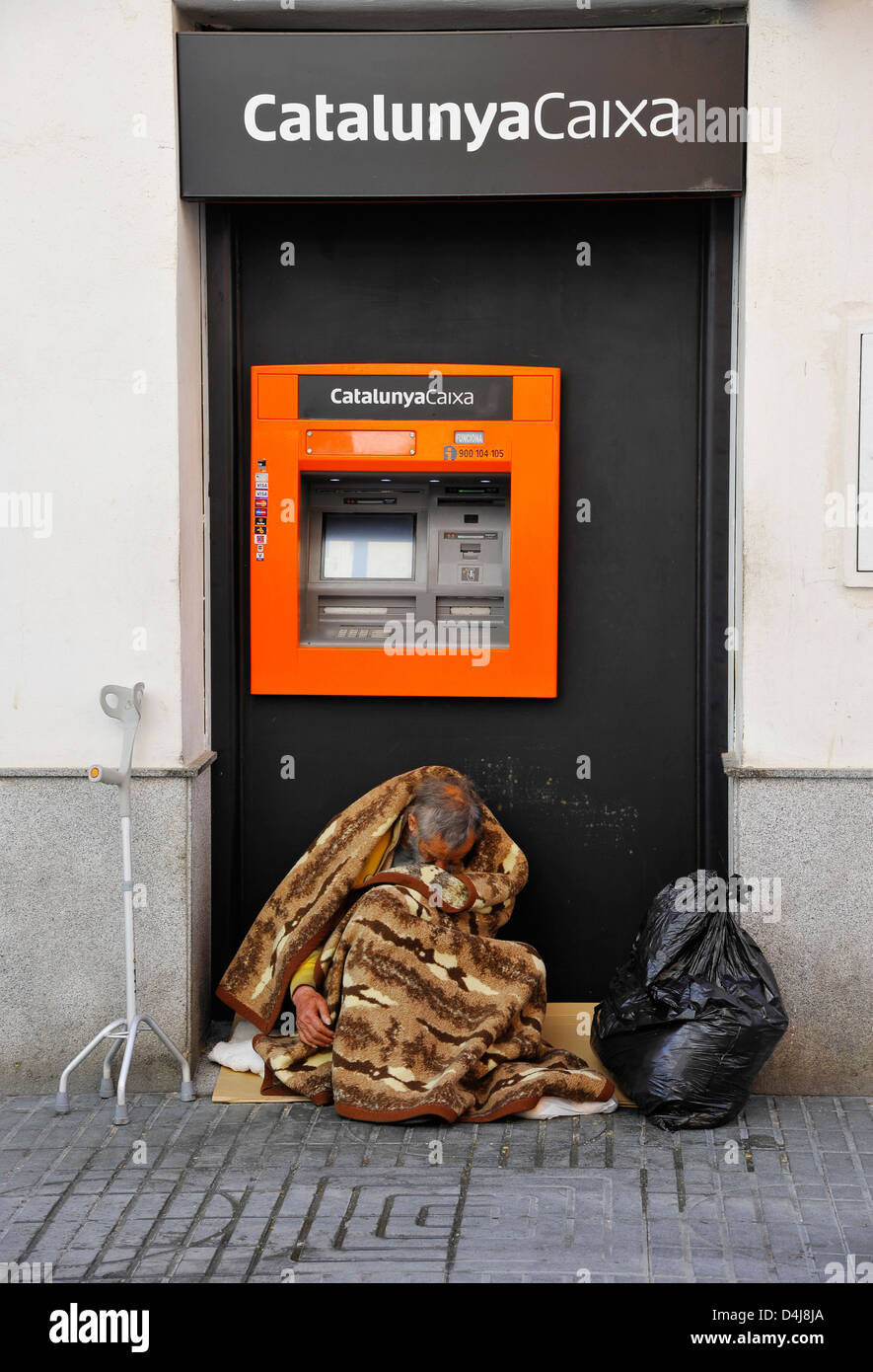 Hombre sin hogar envuelto en una manta se sienta en frente de un cajero automático del banco español 'CatalunyaCaixa' en España Foto de stock