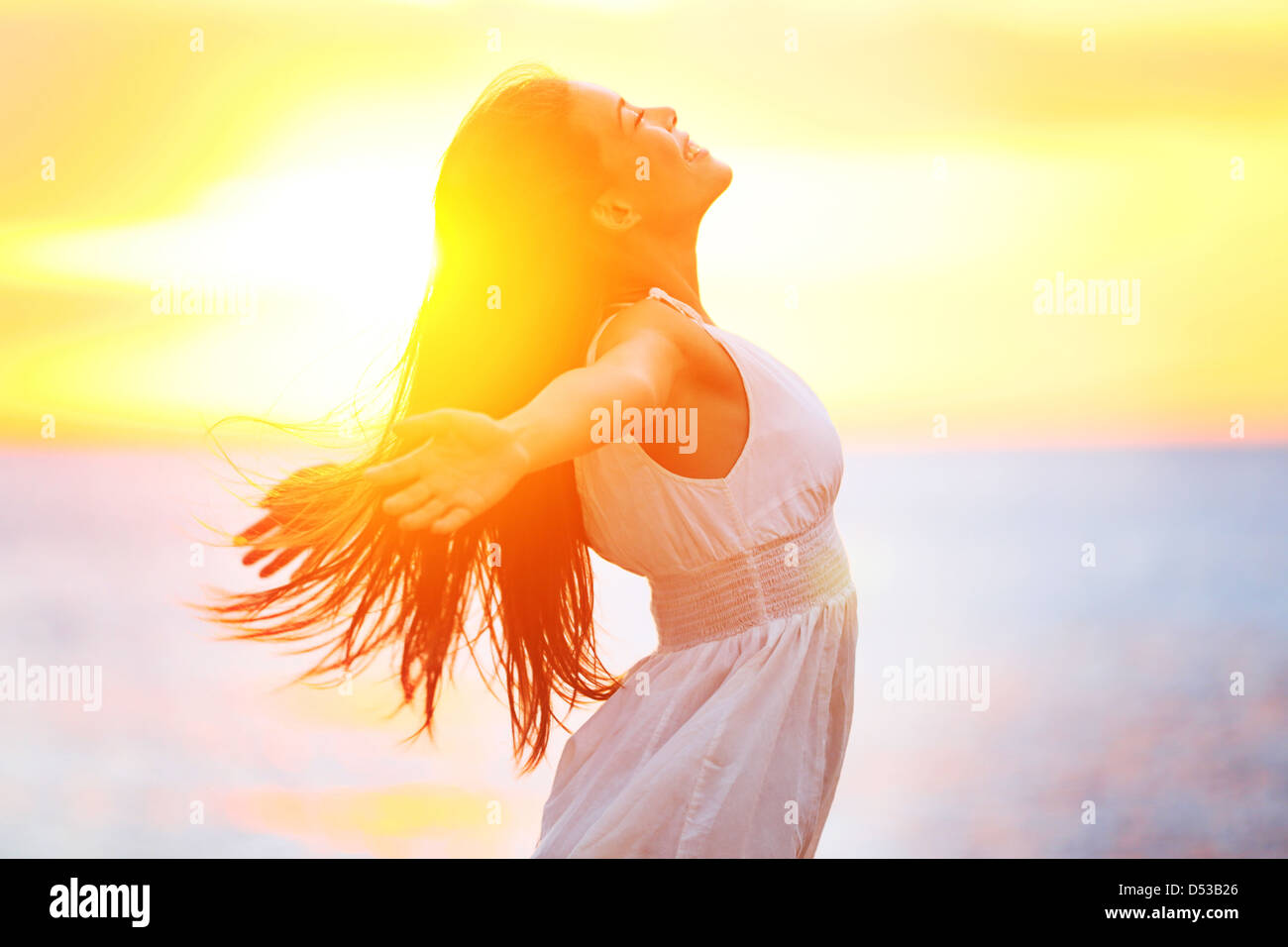 Vista lateral de la feliz joven mujer de raza mixta en vestido blanco con sus brazos extendidos disfrutando de la paz y la serenidad de golden sunet Foto de stock