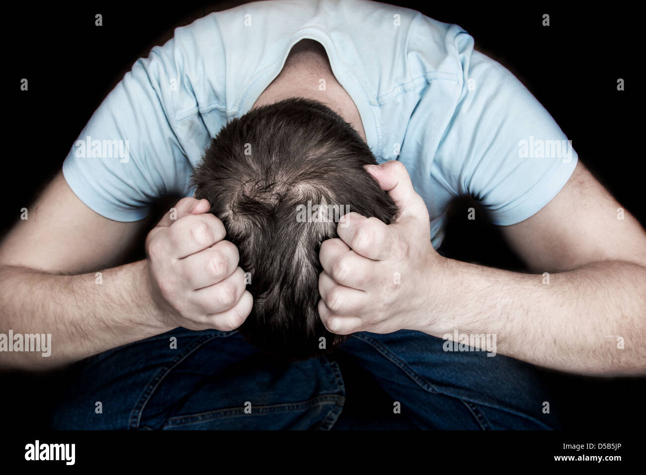 Hombre sujetando su cabeza en sus manos, sentado en el suelo sobre fondo negro. La desesperación, la depresión, la desesperanza, el concepto. Foto de stock