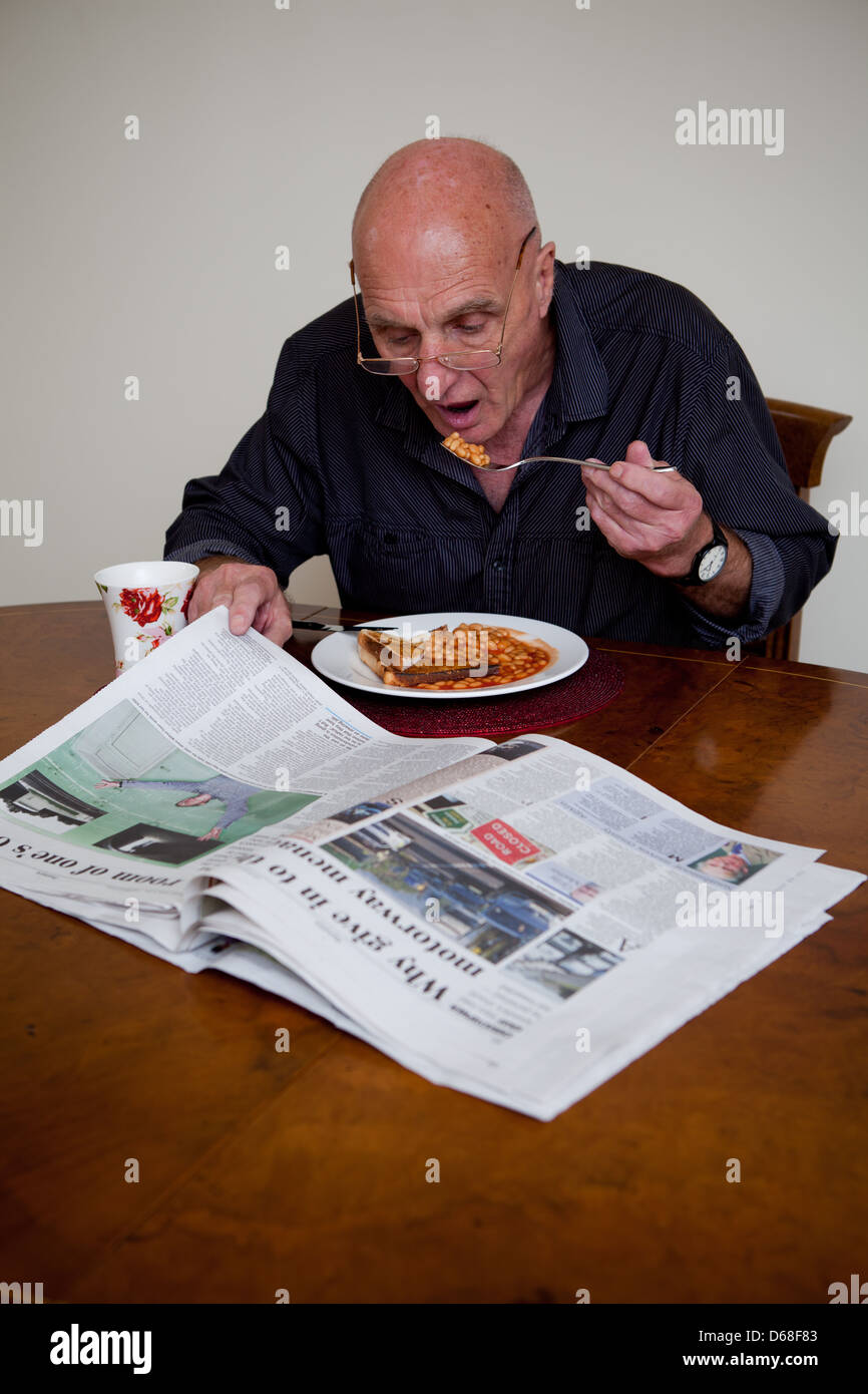 Anciano sentado a la mesa, comer frijoles sobre pan tostado y leyendo un periódico. Foto de stock
